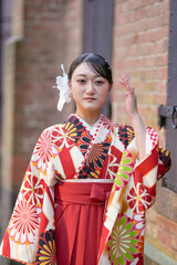 A Japanese woman in her 20s, wearing a red hakama, a staple for Japanese university graduates, stands in front of a brick building.