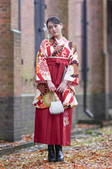 A Japanese woman in her 20s stands on a path filled with autumn leaves in front of a traditional Japanese brick building, wearing a red kimono (hakama), a staple of Japanese university graduates.