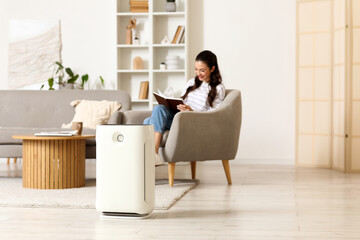 Modern air purifier on floor against woman reading book in armchair at home
