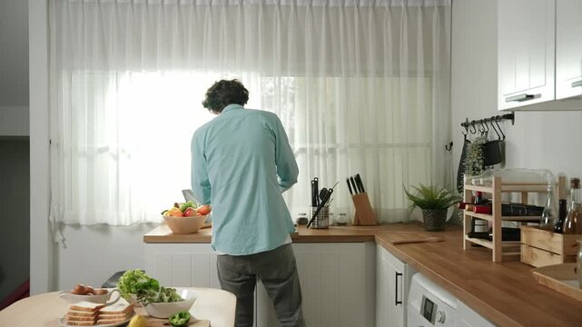 Happy father move to music while cooking food and prepare lunch at modern kitchen. Active dad dancing to song while making healthy food or salad surrounded with vegetable and kitchenware. Pedagogy.