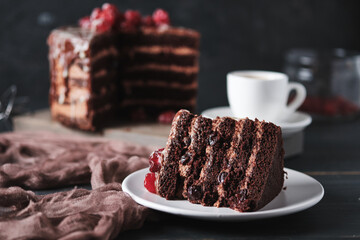 A slice of cake with chocolate and cherry on a plate. A cup of espresso and a cake in the background.