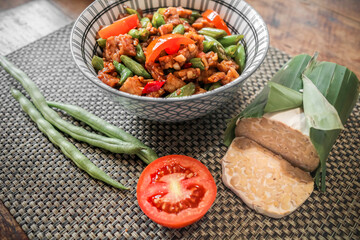 Delicious Indonesian lunch menu of stir fry tempeh and string beans served in a bowl on dining table with raw ingredients on the side