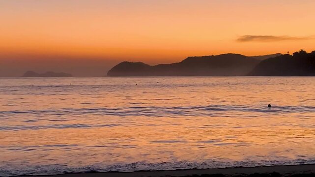 Final de tarde com um tom bem dourado ap&oacute;s o p&ocirc;r do sol e um mar com ondas bem pequenas quebrando na praia. Ao fundo, &agrave; esquerda, uma ilha famosa do Litoral Norte chamada As Ilhas.