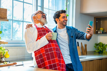 Indian Asian Young son shares smartphone content with senior father enjoying coffee in the kitchen