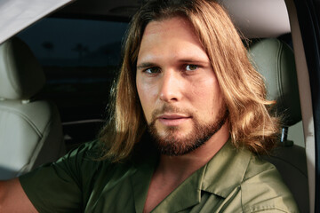 Man with long hair sitting in a car, looking confidently at the camera with a calm expression The...