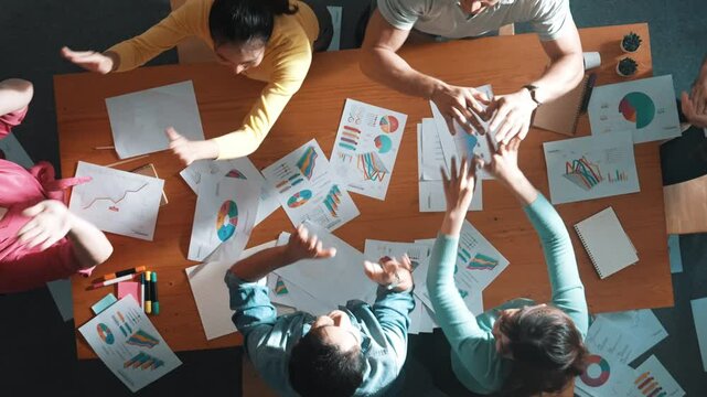 Top view of smart people throw paper and giving high five at meeting table with financial document and stock market analysis paper scattering around. Aerial view of team celebrate project. Symposium.