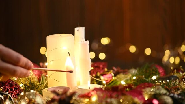 Close up hand lights festive Christmas candles with a match. Burning white candles and Christmas decoration wreath on wooden background.