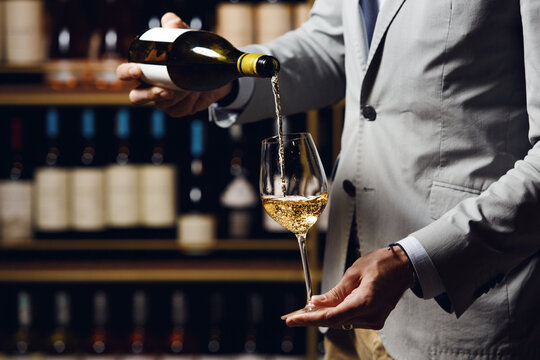 Close up indoor portrait of male sommelier pouring white wine into long-stemmed wineglass, free copy space.