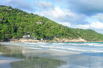 Famous Haad Rin Beach in Koh Phangan with white sandy shoreline, rolling waves, and beachfront resorts nestled against green hillside with granite boulders