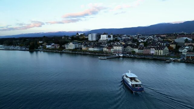 Establishing drone shot of boat arriving in Nyon port with cityscape at sunrise in Canton of Vaud Switzerland
