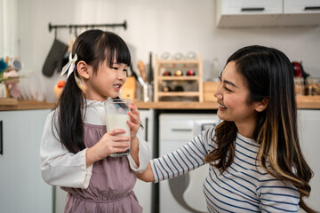 Asian little daughter drinking a glass of milk with mother in kitchen. 