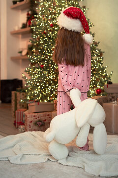 A girl in pajamas and a Santa Claus hat with a toy in her hands looks at a decorated Christmas tree with many gifts