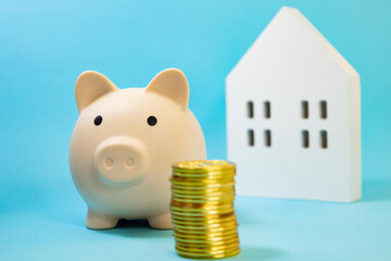 Piggy bank next to stacked gold coins with a white wooden house out of focus behind it. on a blue...
