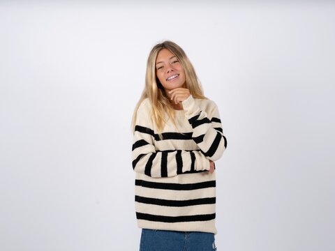 Optimistic Beautiful teen girl wearing striped sweater keeps hands partly crossed and hand under chin, looks at camera with pleasure.