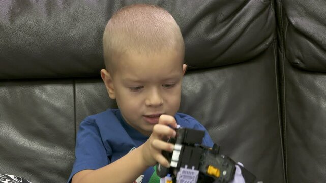 Child playing with new toy sitting on sofa at home. Concentrated face of little boy holding transformer robot toy. Three year old child examining new toy comments on his actions.