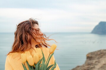 Obraz premium Portrait of a happy woman with hair flying in the wind against the backdrop of mountains and sea. Holding a bouquet of yellow tulips in her hands, wearing a yellow sweater