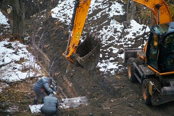 A backhoe loader lays construction slabs, a crew of workers repairs a pipeline using the backhoe...
