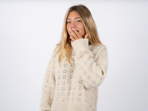 Beautiful teen girl wearing white sweater covers mouth and looks with wonder at camera, cannot believe unexpected rumors.
