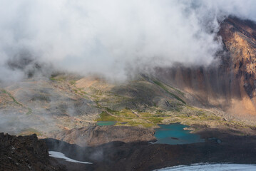 Misty aerial top view to green rocky hilly valley with two turquoise mountain lakes under low...