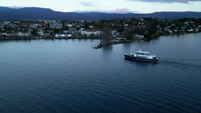 Orbit drone shot of boat sailing to Nyon port at sunrise with cityscape in Canton of Vaud Switzerland