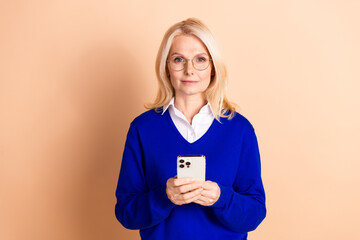 Elegant mature businesswoman with blond hair and glasses holding a smartphone against beige...