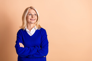 Confident elderly woman in blue jumper posing with arms crossed against beige background, exuding elegance and style.