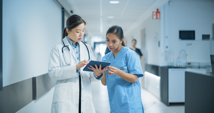 Two Young Asian Female Medical Professionals Standing in a Bright Hospital Corridor with Other People, Engaged in a Serious Discussion While the Doctor Reviews Patient Details on a Tablet Computer - Powered by Adobe
