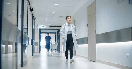 Fototapeta premium Asian Healthcare Professional Walking to Her Examination Room, Reception Desk, ICU Unit in a Modern Bright Hospital Corridor. Doctor Walking with Her Hands in Pocket