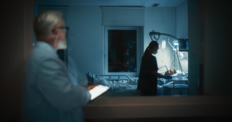 Senior Doctor Standing in the Dark Hallway Outside a Hospital Intensive Care Ward, Focused on a Tablet Computer while Observing a Nurse Attending to a Patient Resting in the Bed Beyond the Glass