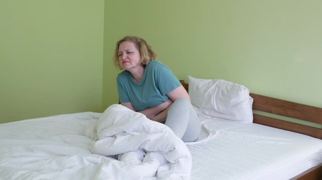A middle-aged woman sits on the bed in the bedroom and suffers from abdominal pain, diarrhea, gastritis, colitis, gases, menstruation