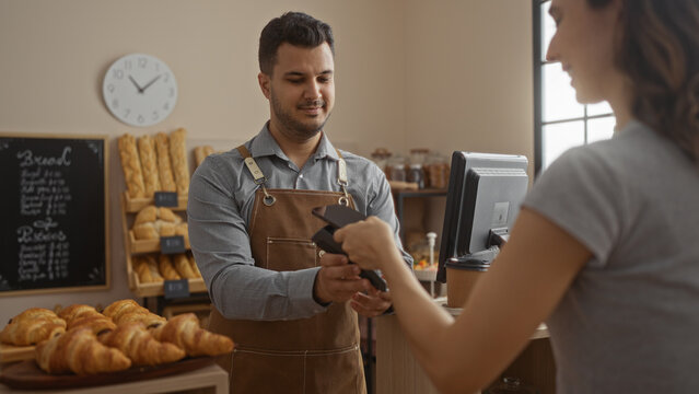Woman paying with card to a smiling man in a bakery filled with croissants and baguettes