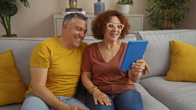 Middle-aged couple using tablet together in cozy living room, man and woman sitting on sofa smiling and engaged