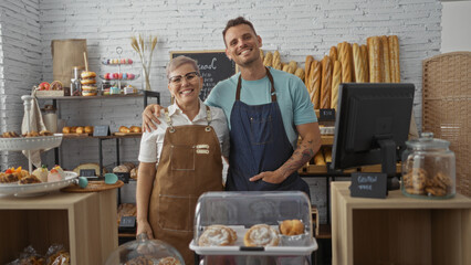 Woman and man bakers wearing aprons smiling and posing together in a cozy bakery interior filled with breads and pastries.
