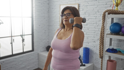 Middle-aged hispanic woman lifting dumbbell in a gym setting, wearing exercise clothes, performing strength training indoors with sports equipment in the background.