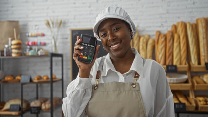 African american woman working in a bakery indoors, smiling and holding a payment terminal,...
