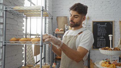 Handsome young hispanic man with a beard arranging baked goods on a shelf in a cozy bakery shop