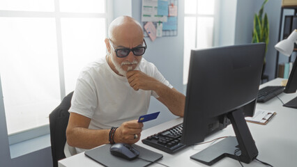 Fototapeta premium Mature, grey-haired man sitting in an office holds a blue credit card while working on a computer, concentrating on an online financial transaction.