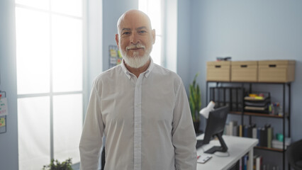 Middle-aged man with grey hair and beard smiling in a modern indoor office setting with shelves and...