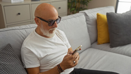 Man with grey beard and glasses sitting on a grey couch in a home living room, engrossed in his...