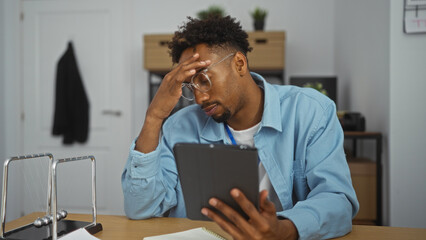 A young african american man with a beard looks stressed while holding a tablet in an office...