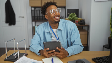 Fototapeta premium Handsome african american man with a beard smiling in an office environment holding a tablet, surrounded by paperwork and office supplies.