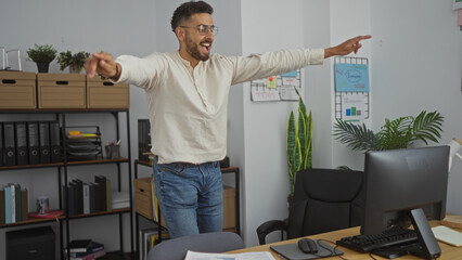 Happy young hispanic man celebrating in a modern office, expressing joy and excitement with arms...