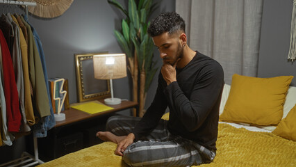 Handsome young hispanic man sitting thoughtfully on a bed in a stylish bedroom with yellow accents...