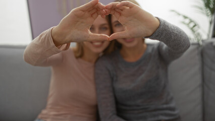 Sisters posing happily on a couch, forming a heart with their hands in a cozy living room, showcasing love, connection, and family unity in an intimate indoor setting.