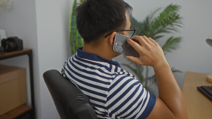 A young chinese man in an office setting is talking on a smartphone while seated at a desk indoors, wearing a striped shirt, and surrounded by various office supplies and plants.