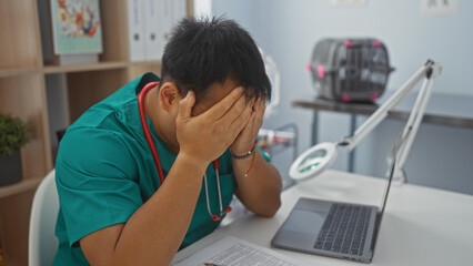 Young asian man in a green uniform looks stressed at work in a veterinary clinic, with a laptop and...