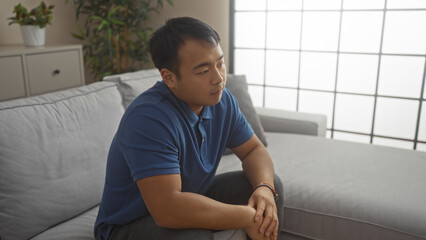 Fototapeta premium A young chinese man sits thoughtfully in a stylish living room, showcasing a modern interior with natural light, plants, and comfortable furniture, creating a serene and contemplative atmosphere.