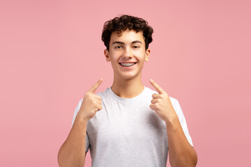Excited, smiling boy, teenager pointing fingers at his teeth with braces isolated on pink background