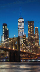 Fototapeta premium The photo captures the Brooklyn Bridge at night from Brooklyn, with illuminated city lights reflecting on the calm water. The skyline, a modern skyscraper, and the serene park enhance the scene.