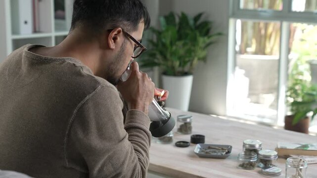 Asian man smoking cannabis with a bong in a living room, with cannabis equipment and accessories on the table.